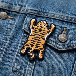 May include: An embroidered patch shaped like a tiger with orange fur, black stripes, and a white belly. The tiger is depicted in a flat, spread-out position with its limbs extended. The patch is attached to the front of a blue denim jacket.