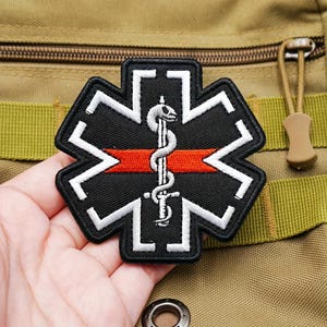 May include: A black embroidered patch in the shape of a six-pointed star of life. The design features a white outline, a horizontal red stripe across the center, and a white serpent coiled around a vertical staff in the middle of the patch.
