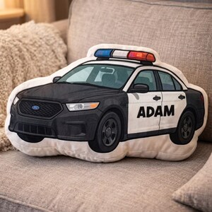 May include: A plush police car pillow in black, white, and gray, with a blue and red light bar. The car features the name "ADAM" on the side. The pillow is designed to look like a police cruiser.