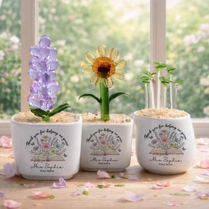 Könnte beinhalten: Drei weiße Keramik-Töpfe mit Glasblumen und Pflanzen. Jeder Topf zeigt eine Blumenillustration, ein offenes Buch und den Text "Thank you for helping me grow", "Mrs. Sophia" und "From Byron" in schwarzer Schreibschrift.