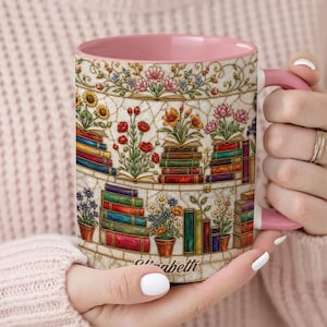 Könnte beinhalten: Ein Keramikbecher mit rosafarbenem Innenraum und Henkel, verziert mit einer Illustration von Bücherstapeln und Topfblumen im Buntglas-Stil. Der Name Elizabeth ist in Schreibschrift am unteren Rand des Becherdesigns aufgedruckt.