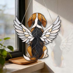 Peut inclure: Un attrape-soleil en vitrail représentant un chien vu de dos avec de grandes ailes blanches. Le chien présente un pelage tricolore composé de segments de verre noir, marron et blanc. L'objet est suspendu par une chaîne en métal argenté.