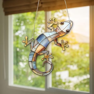 Op de afbeelding: Een gebrandschilderde glazen gecko-zonnevanger, hangend aan een zilveren ketting. De gecko heeft een mozaïekontwerp met tinten blauw, bruin en goud. De voeten van de gecko zijn goudkleurig. De achtergrond is een vage weergave van een raam en groen.