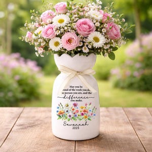 Könnte beinhalten: Eine weiße Keramikvase mit gewelltem Rand und cremefarbenem Band, gefüllt mit rosa Rosen und weißen Gänseblümchen. Die Vase trägt den Text: May you be proud of the work you do, the person you are, and the difference you make. Savannah 2025.