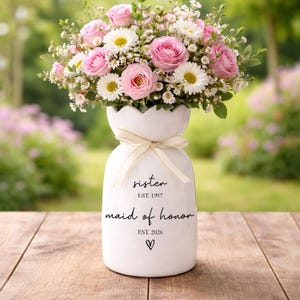 Könnte beinhalten: Eine weiße Keramikvase mit gewelltem Rand und einem cremefarbenen Band um den Hals. Die Vase trägt den schwarzen Schriftzug "sister EST. 1997", "maid of honor EST. 2026" sowie ein kleines Herzsymbol und ist mit rosa Rosen und weißen Margeriten gefüllt.