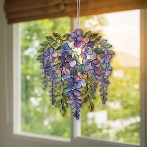 Könnte beinhalten: Eine hängende Fensterdekoration aus Buntglas mit einem Cluster aus lila Blauregenblüten und grünen Blättern. Das florale Design wird von einer silbernen Kette gehalten und eignet sich als dekorativer Akzent für ein Fenster oder eine Wand. Die Blüten sind in Lila- und Blautönen gehalten.