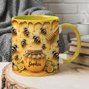 Op de afbeelding: Een keramische mok met een gele binnenkant en handvat, voorzien van een honingthema met meerdere bijen, een houten honingdipper en een centrale illustratie van een honingpot. De pot heeft een deksel in jute-stijl en de naam Sophia in zwart.