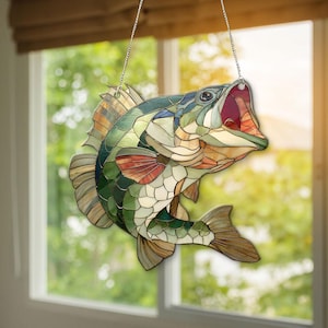 May include: A stained glass suncatcher shaped like a largemouth bass fish, featuring a mosaic of green, tan, and red glass segments held together by dark metal lines, suspended by a thin silver chain against a bright window background.