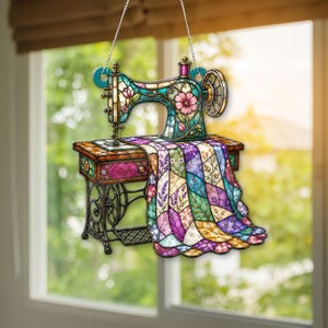 Puede incluir: Un adorno colgante con forma de máquina de coser vintage con estética de vidriera. La máquina presenta motivos florales en turquesa, rosa y morado, con una colcha de retazos multicolor drapeada que cuelga de la mesa de costura.