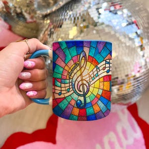 Könnte beinhalten: Ein Keramikbecher mit blauem Henkel und einem farbenfrohen Buntglas-Muster aus einem Violinschlüssel und Noten. Der Name Amanda ist in schwarzer Schreibschrift in der Mitte des Violinschlüssels vor einem leuchtenden Mosaikhintergrund gedruckt.