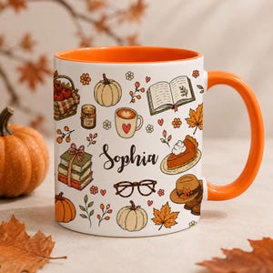 Peut inclure: Un mug en céramique blanc avec un intérieur et une anse orange, orné d'un motif automnal répétitif composé de citrouilles, de livres, d'une tasse de café, de lunettes, d'un chapeau et d'une part de tarte. Le prénom Sophia est écrit en noir.