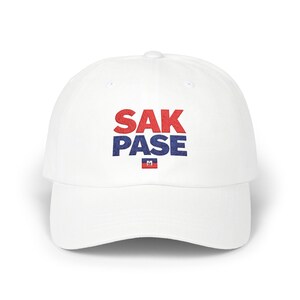 May include: White baseball cap with the text "SAK PASE" in red and blue block letters. A small Haitian flag is below the text. The cap has a curved brim and a rounded crown.