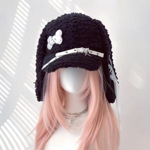 Pode incluir: Um chapéu preto texturizado com uma aba branca e detalhes decorativos, incluindo um laço e uma tira com detalhes metálicos. O chapéu tem longas extensões semelhantes a orelhas e é exibido em uma cabeça de manequim com cabelo rosa.