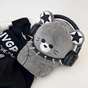 Pode incluir: Boné de beisebol cinzento com um design de rosto de urso, detalhes de estrelas pretas e uma coleira com pontas. Os fones de ouvido pretos estão colocados no boné. Uma camisa preta com texto branco é visível.