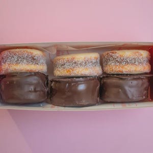 May include: A box containing six alfajores, a traditional South American sweet treat. The alfajores have a chocolate base, a dulce de leche filling, and a coconut dusting. The box is pink, set against a pink backdrop.