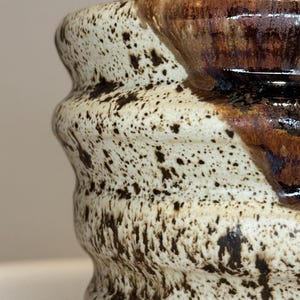 Puede incluir: Primer plano de una taza de cerámica con un exterior moteado blanquecino y un borde marrón oscuro. La taza tiene un diseño ondulado único y un acabado brillante. El patrón moteado es marrón oscuro.