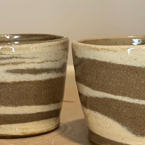 May include: Two ceramic cups with a striped design. The cups have a cylindrical shape with alternating bands of brown and off-white. The cups are handmade and have a rustic, earthy aesthetic.