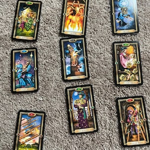 May include: Nine tarot cards with black borders are displayed on a neutral surface. The cards feature colorful illustrations and titles such as "The Devil," "The Star," and "King of Pentacles." The cards are from a tarot deck.