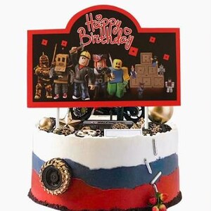 Puede incluir: Un adorno para tarta redondo con un cartel rectangular que dice Happy Birthday y un grupo de figuras estilo bloque. La tarta est&aacute; decorada con glaseado blanco, azul y rojo, un neum&aacute;tico negro y peque&ntilde;as piezas met&aacute;licas de color plateado.