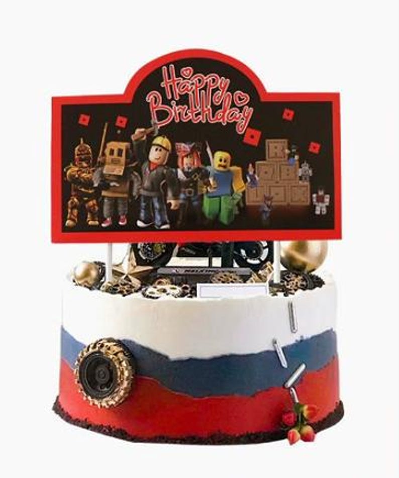 Puede incluir: Un adorno para tarta redondo con un cartel rectangular que dice Happy Birthday y un grupo de figuras estilo bloque. La tarta est&aacute; decorada con glaseado blanco, azul y rojo, un neum&aacute;tico negro y peque&ntilde;as piezas met&aacute;licas de color plateado.
