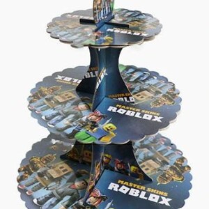 Puede incluir: Un soporte para cupcakes de cart&oacute;n de tres niveles con un tema azul y azul oscuro y gr&aacute;ficos de Roblox Master Skins. Los niveles circulares tienen bordes festoneados y una estructura central con el texto Master Skins Roblox impreso.