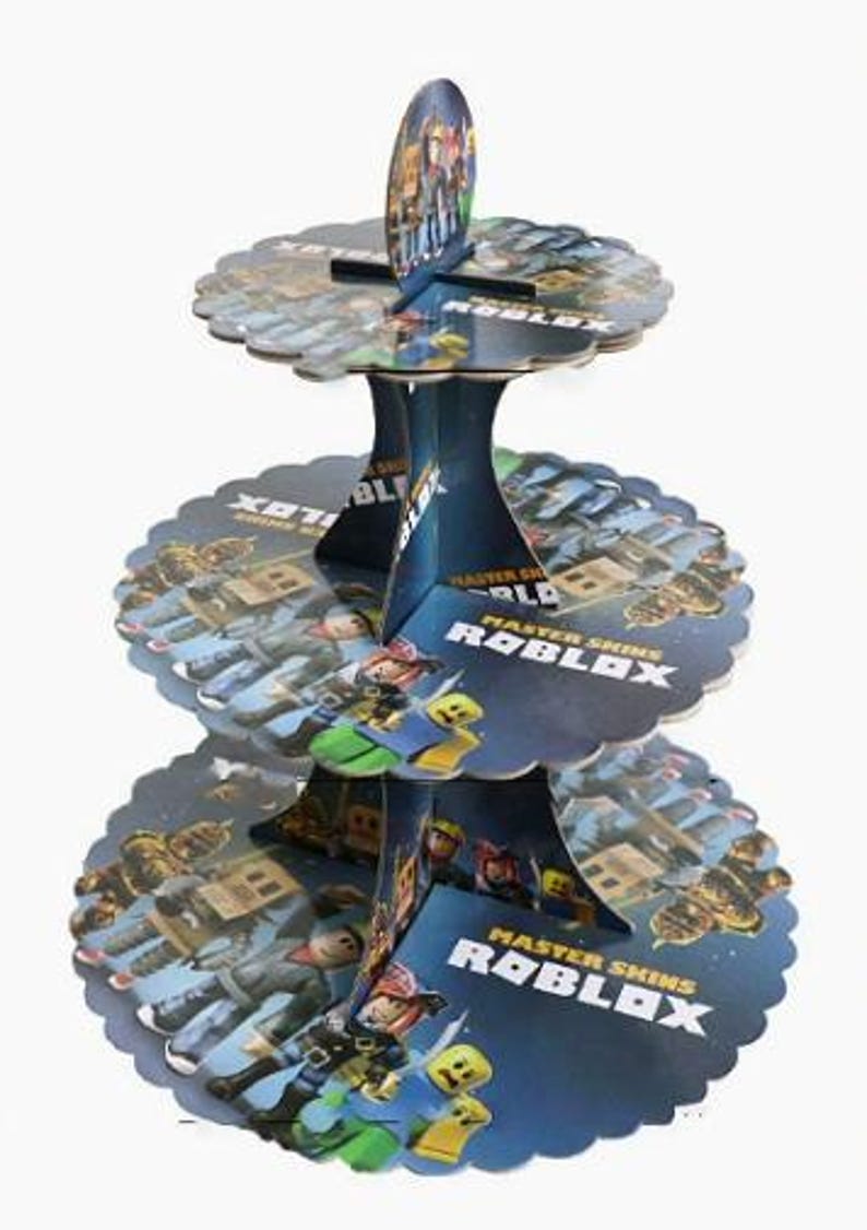 Puede incluir: Un soporte para cupcakes de cart&oacute;n de tres niveles con un tema azul y azul oscuro y gr&aacute;ficos de Roblox Master Skins. Los niveles circulares tienen bordes festoneados y una estructura central con el texto Master Skins Roblox impreso.