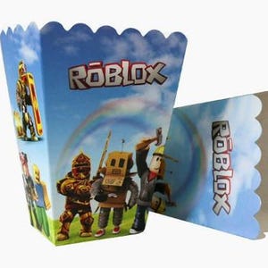 Puede incluir: Una caja de palomitas de papel con un fondo de cielo azul, un arco&iacute;ris y el logotipo de Roblox. La caja muestra varias figuras de personajes del juego, incluido un personaje tipo robot y una figura con un casco amarillo.