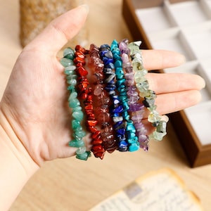 Può includere: Una mano mostra sette fili di braccialetti con schegge di pietre preziose in vari colori. Le pietre preziose includono verde, rosso, marrone, blu, viola e turchese. I braccialetti sono infilati, mostrando la bellezza naturale delle pietre.