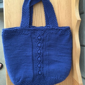 May include: A blue knitted tote bag with a cable knit design and a decorative stitch detail. The bag has two straps.