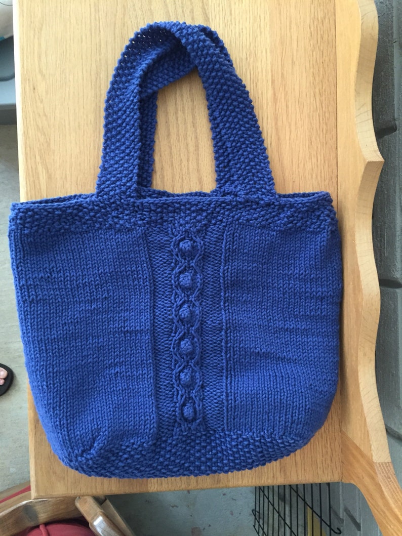 May include: A blue knitted tote bag with a cable knit design and a decorative stitch detail. The bag has two straps.