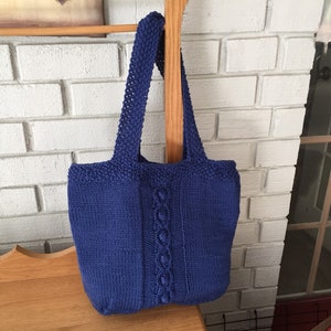 May include: A blue knitted tote bag with a cable knit design. The bag has two straps and is hanging on a wooden dowel.