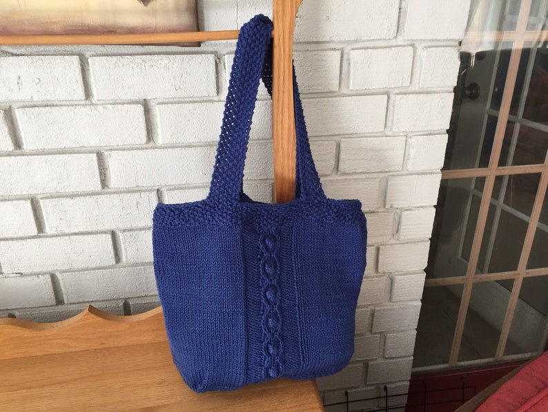 May include: A blue knitted tote bag with a cable knit design. The bag has two straps and is hanging on a wooden dowel.