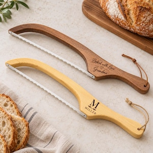 Könnte beinhalten: Zwei Brotmesser aus Holz mit gezackten Metallklingen und Lederschlaufen zum Aufhängen. Das dunkle Holz ist mit "Baked with Love Grandma" graviert, das helle Holz trägt den Buchstaben "M" und den Namen "Miller".