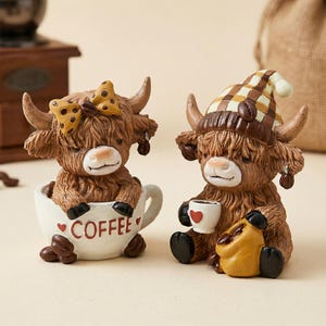 May include: Two Highland cow figurines. One cow is in a coffee cup with the word "COFFEE" on the cup. The other cow is wearing a hat and holding a coffee cup and a bag of coffee beans. The figurines are brown and white.