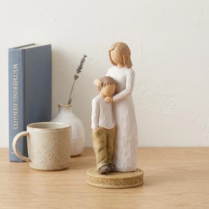 May include: A carved figurine of a mother and child. The mother is in a white dress, and the child is in a white shirt and tan pants. The figurine is on a decorative base. A mug and a book titled "Wuthering Heights" are in the background.
