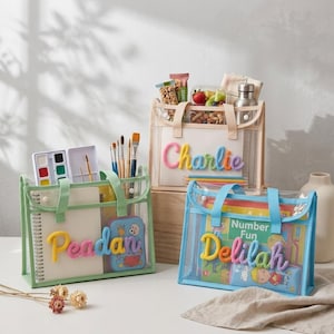 May include: Three rectangular clear plastic tote bags with handles, each featuring a personalized name in colorful yarn lettering on the front. The bags contain various items including notebooks, paint sets, art brushes, and books.
