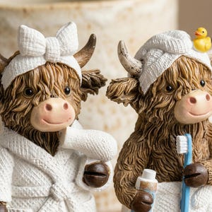 May include: Two Highland cow figurines, one with a white bow headband and robe, the other with a white cap, a rubber duck, and holding a toothbrush and toothpaste. The cows have brown fur, pink noses, and are smiling.