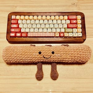 May include: A wooden-framed keyboard with colorful keycaps, including red, white, and beige. Below, a crocheted brown wrist rest shaped like a smiling bread roll with small legs. The keyboard has a variety of symbols and letters.