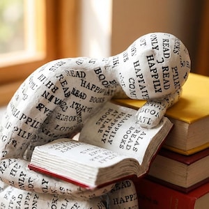 Könnte beinhalten: Eine weiße Skulptur einer lesenden Person, auf der die Worte „Read Happily“ und andere buchbezogene Phrasen aufgedruckt sind. Die Skulptur ruht auf einem Stapel von Büchern mit roten und gelben Einbänden.
