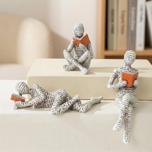 以下が含まれることがあります： 読書をする人々の装飾的な置物3体。 白地に黒い文字模様で、オレンジ色の本を持っています。 一体は横たわり、もう一体はあぐらをかき、もう一体は足を伸ばして座っています。