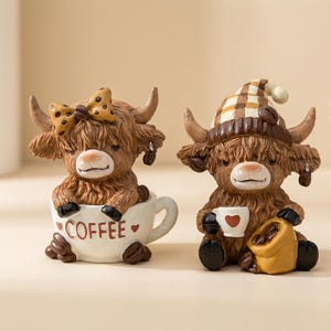 May include: Two Highland cow figurines. One cow is in a coffee cup with the word "COFFEE" on it, and the other cow is wearing a hat and holding a coffee cup. Both figurines are brown with horns and coffee bean accents.