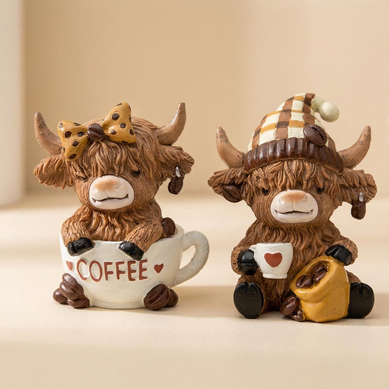 May include: Two Highland cow figurines. One cow is in a coffee cup with the word "COFFEE" on it, and the other cow is wearing a hat and holding a coffee cup. Both figurines are brown with horns and coffee bean accents.