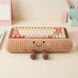 May include: A wooden-framed keyboard with a variety of pastel-colored keys, including pink, white, and yellow. A crocheted wrist rest in a light brown color with a smiling face and small crocheted feet is positioned in front of the keyboard.