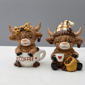 May include: Two Highland cow figurines. One cow is in a coffee cup with the word "COFFEE" on it, and the other cow is wearing a nightcap and holding a coffee cup and a bag of coffee beans. The figurines are brown and tan.