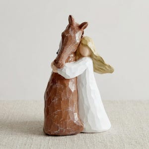 May include: A sculpted figurine featuring a person with long blonde hair wearing a white dress, embracing a brown horse. The piece has a carved, textured appearance with visible facets, showing the person hugging the horse's neck.