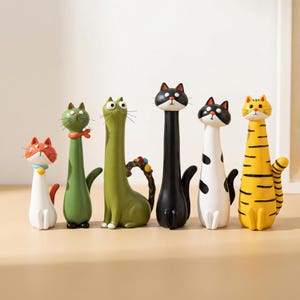 Könnte beinhalten: Eine Sammlung von sechs skurrilen Katzenfiguren in verschiedenen Farben und Mustern. Die Katzen haben längliche Körper und Hälse mit Details wie Streifen, Flecken und Accessoires. Die Figuren sind aus Keramik oder einem ähnlichen Material gefertigt.