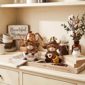 May include: Two Highland cow figurines, one in a coffee cup with "COFFEE" text, the other holding a coffee cup and a bag of beans. They sit on a wooden tray with coffee beans, near a sign that says "Brewtiful Mornings".