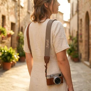 May include: A camera strap made of a textured beige fabric with brown leather accents and the name Sarah embroidered in cursive script. The strap is attached to a brown and silver compact camera, featuring a lens and a shutter button on top.