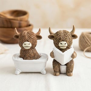 May include: Two Highland cow figurines. One is in a white bathtub, the other is reading a white book. Both have brown fur, horns, and white faces. The bathtub figurine is surrounded by white material. The book is white.