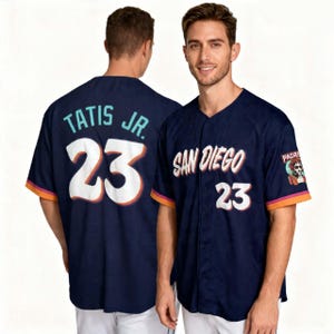 以下が含まれることがあります： 紺色の野球ユニフォーム。白文字で「San Diego」と「23」と書かれています。袖にはオレンジとピンクのトリムがあり、スカルデザインのパッチが付いています。背中には「Tatis Jr.」と数字の「23」が表示されています。
